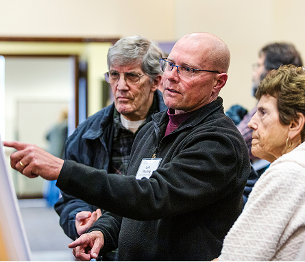 A staff member at a public meeting explaining project information on a display board to several attendees who are standing close and following along.