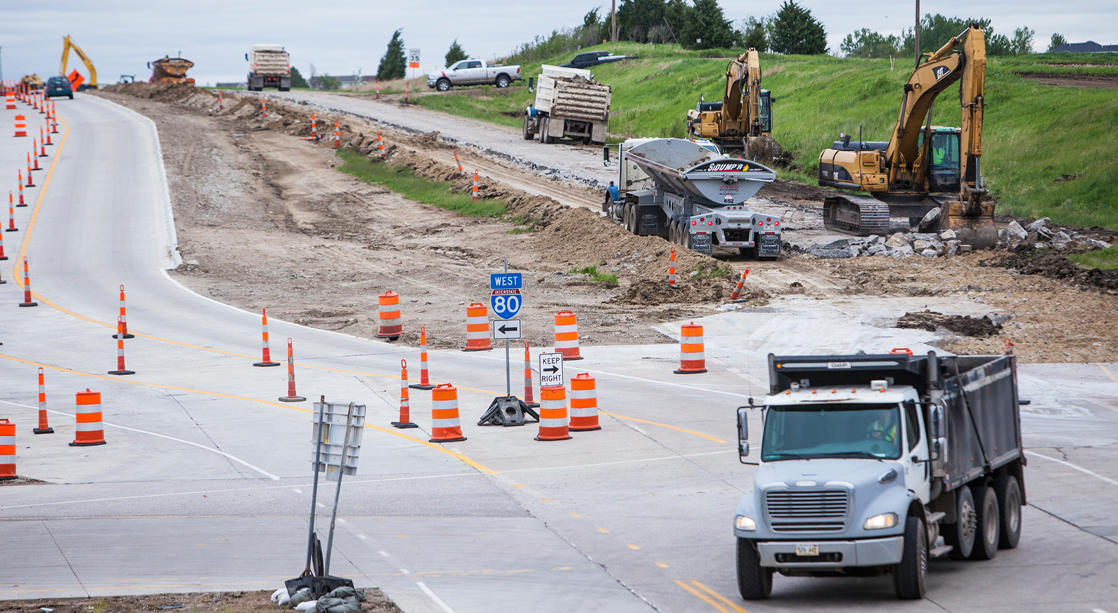 Active roadway construction along the US 75 corridor, showing traffic redirected through a work zone marked by orange barrels and cones. Construction vehicles and heavy equipment are working beside partially completed pavement.